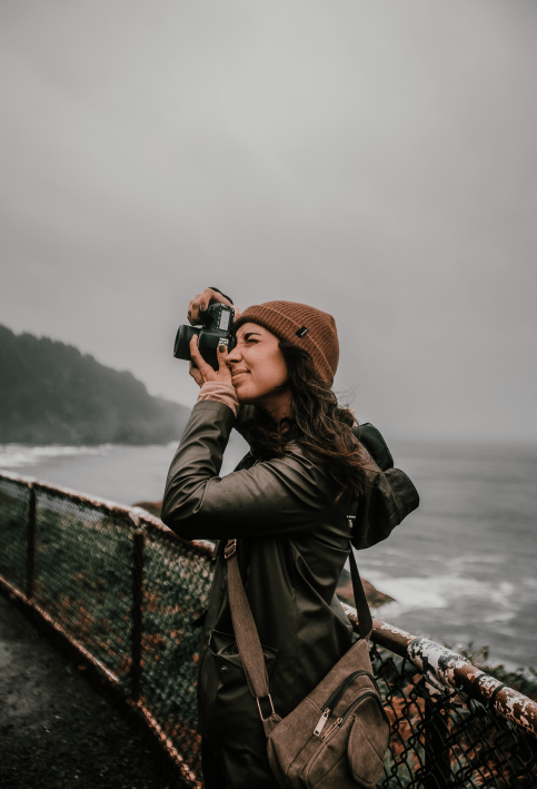 a woman in a green rainjacket near the ocean taking photographs with a dslr camera on a rainy day.