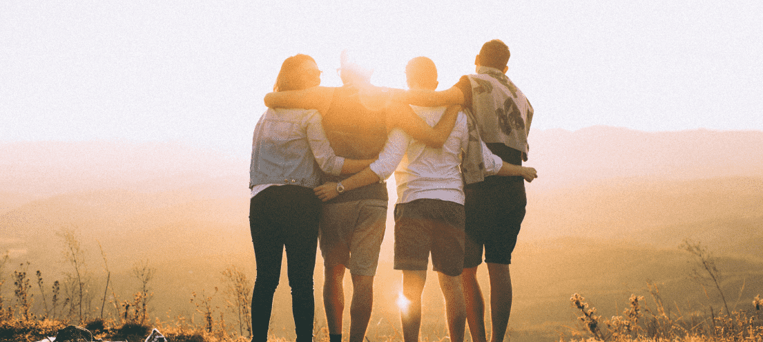 a group of four people standing on a mountain top, holding eachother cheerfully.