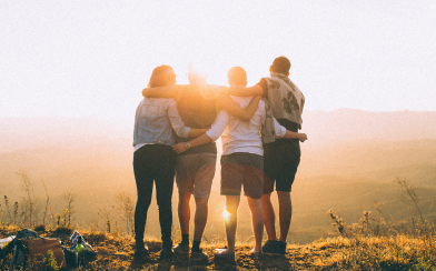 a group of four people standing on a mountain top, holding eachother cheerfully.
