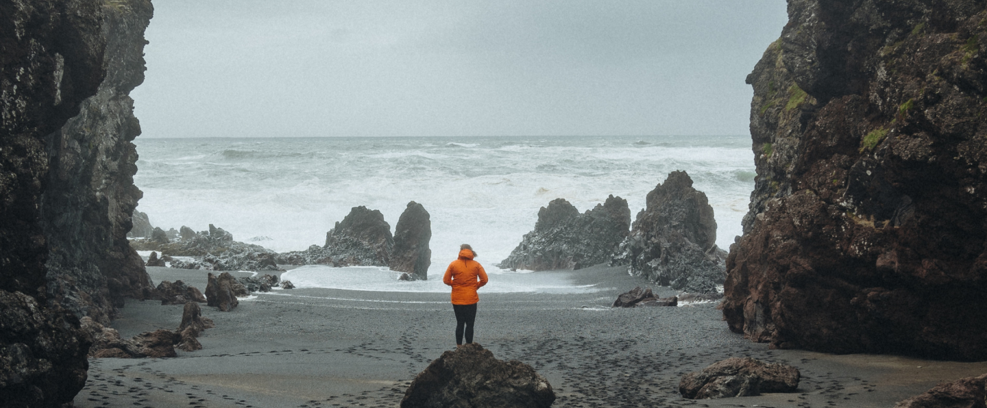 A woman in a rainjacket walking on a rainy beach where the ocean is storming